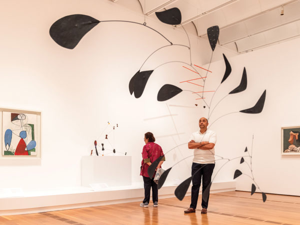 A visitor stands in a gallery looking up at a large black and red mobile while another visitor behind him looks at a statue on a pedestal.