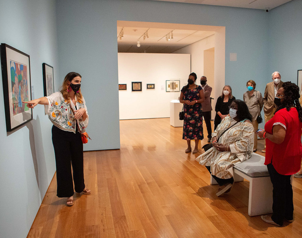 A woman points to a framed artwork on the wall and talks to a group of people in a gallery.