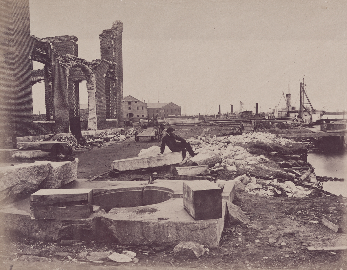 A man sits atop a slab of stone looking toward docked boats, the ruins of a large brick building behind him.