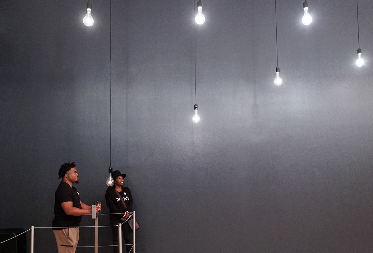 Two visitors in Rafael Lozano-Hemmer's Pulse Room. One person holds the metal sensors to have their pulse recorded, while the two people look at the lightbulbs hanging from the ceiling.