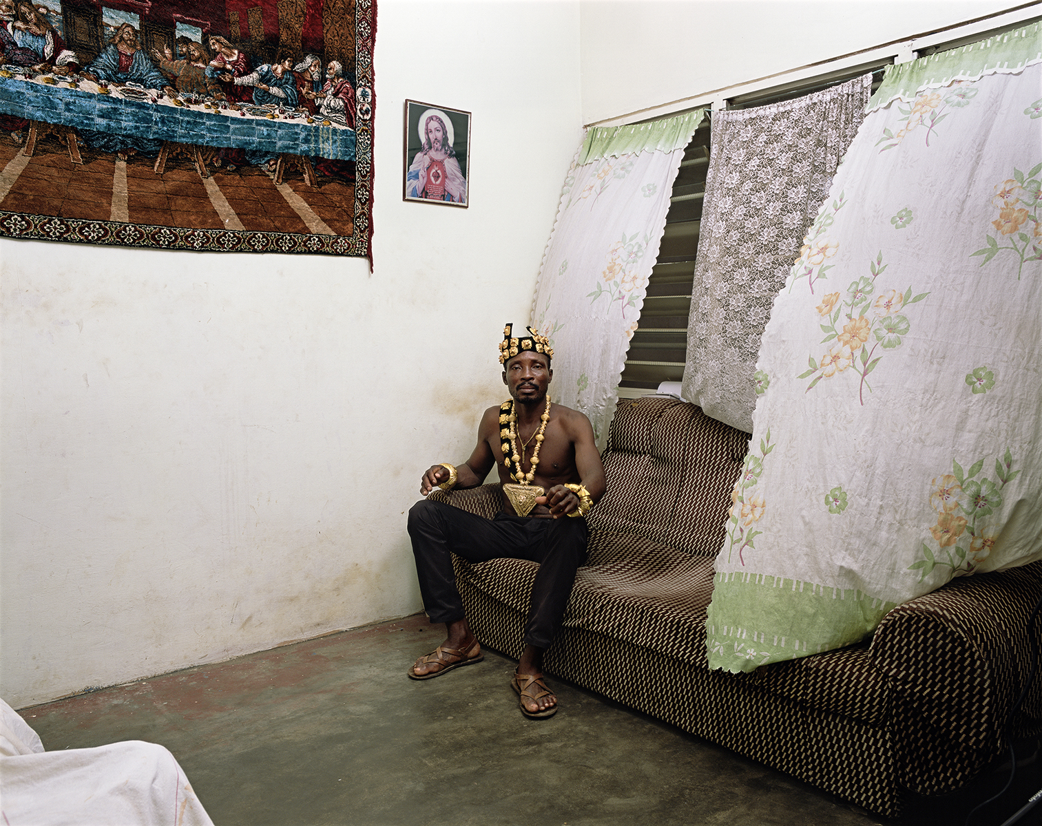 Photograph of a man wearing an ornate gold necklace and headpiece seated on a sofa in front of billowing curtains. Religious images hang on the wall beside him.