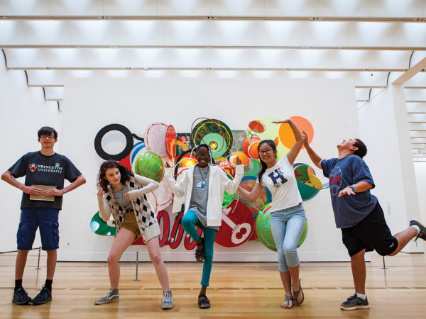 Five children hold different poses in front of an three dimensional artwork.