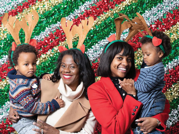 Two women, each holding a small child pose for the camera in front of a backdrop of red, green, gold, and silver bows.